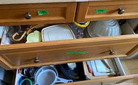 Open drawers showing strainers, fry pan, cutting boards, glass tumblers, lids, tins, wicker basket