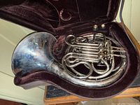 French horn resting in open maroon-lined molded hard case with two mouthpieces visible in lid compartment