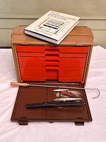 Open tackle box showing red drawers, knives, lures, rod-building book on lid
