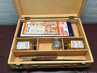 Case open showing contents: mediums, palette cup, brushes, booklet, paint tubes beneath divider