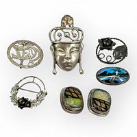 Six assorted silver pins, brooches, and clip-on earrings arranged on a white background to display varied designs and colors.