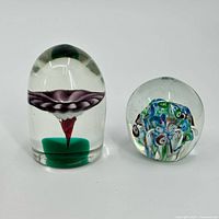 Two glass paperweights shown side by side on white background: one oblong with a purple flower, the other round with blue and green millefiori floral design.