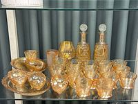 Full lot of marigold glass items displayed on a glass shelf including two decanters with stoppers, multiple cups, bowls, and trays.
