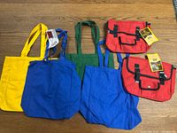 Group of four cotton tote bags in yellow, green, blue shades and two red messenger bags on wooden floor.