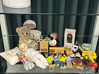 Display of various vintage toys arranged on a reflective surface, including dolls, teddy bear, toy furniture, and accessories.