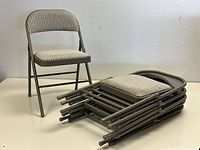 Photo shows one gray folding chair upright and five chairs folded and stacked horizontally on a surface, showcasing cushioned ribbed fabric seats and metal frames.
