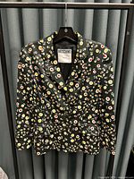 Front view of black Moschino Jeans jacket with white, yellow, and red floral pattern, buttoned front, hanging on hanger.