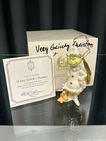 Porcelain Grinch ornament standing beside original Lenox box and certificate of authenticity on reflective surface under lighting.