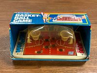 Front angled view of basketball game in original box showing red court, yellow hoops under clear plastic dome and scoring sliders on the sides.