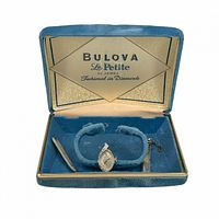 Blue velvet lined open box with gold trim showing Bulova La Petite branding inside the lid and containing the watch band and loose strap pieces.