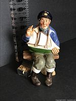 Front view of Royal Doulton Tall Story figurine showing seated sailor with model boat and tackle box, height scale visible