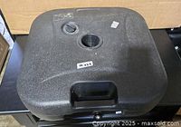 Top view of square black plastic umbrella base showing pole opening, cap, surface texture and handle