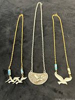 Three necklaces laid out: two gold-tone with bird pendants and blue beads, one silver-tone with pewter bird medallion