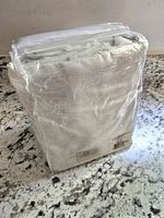 Curtain panel folded and sealed inside original clear plastic packaging, showing folded textured white fabric and attached header for hanging.