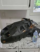 Side view of the large black Batman Tumbler vehicle model on a white granite countertop, showing angular design and golden mechanical details, with water bottle for scale.