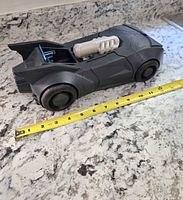 Side view of black Batman Batmobile toy with tape measure showing length approximately 11 inches. Batmobile has blue translucent cockpit and silver engine detail on top.