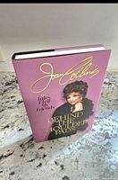 Front cover of Joan Collins Behind the Shoulder Pads book with pink and gold embossed details and image of Joan Collins.