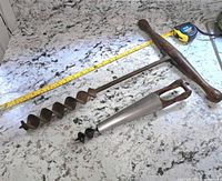 Two hand augers laid on a marble countertop with a measuring tape for scale. The wooden-handled auger is about 14 inches long, metal-handled auger about 8 inches long.