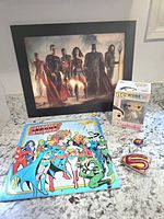 Framed Justice League lenticular print showing Aquaman, Cyborg, Flash, Superman, Wonder Woman, Batman, and other characters, displayed behind a Justice League calendar and collectible items on a granite countertop.
