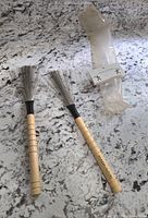 Two new wire drum brushes with wooden handles on a white granite countertop. One brush has brand marking 'LAWN STAKE' on handle. Clear plastic sleeve with label beside brushes.