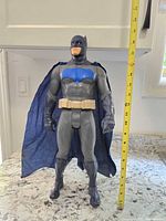 Front view of Batman figure standing next to a measuring tape showing approximately 15 inches tall, with details of suit, bat emblem, and utility belt visible.