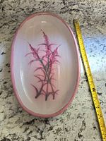 Top-down view of large oval ceramic plate with floral pink design and measuring tape showing length around 16 inches for scale