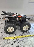 Side view of the black Batman monster truck toy with oversized tires and Batman logo on side, a measuring tape showing scale.