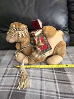 Plush camel toy seated on a surface with decorated fabric saddle and beaded tassels, measuring about 12 inches in length.