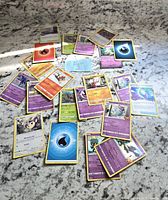 Photo showing approximately 30 assorted loose Pokémon cards spread on a light gray marbled surface. Cards are mixed types and colors, including psychic, fire, water, electric, and grass energy cards, plus multiple different Pokémon character cards.