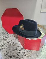 Black fedora hat with grosgrain ribbon band and feather, displayed on original red hexagonal box on a granite countertop.