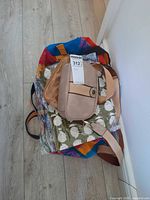 Stack of assorted bags showing beige crossbody, tan pouch, green printed tote in plastic, multicolor tote beneath