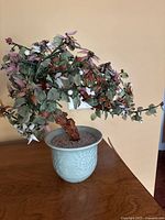Artificial bonsai tree with synthetic leaves and berries in pale blue-green ceramic pot with embossed details, viewed from front.