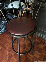Front view of one bar stool showing maroon faux leather seat, dark brown metal frame, circular footrest, and metal backrest with curved top rail