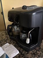 Front view of black DeLonghi Caffè Nabucco machine showing two glass carafes, control panel, and steam wand with milk frother attachment.