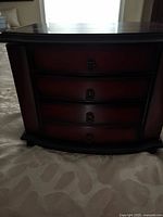 Front view of the dark reddish brown wood jewelry box showing four drawers with ornate metal handles, slightly curved front and polished finish.