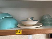 Four Pyrex bowls arranged on a shelf, two stacked light turquoise bowls, two on their sides, one white plate and a white and blue bowl in center.