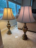 Pair of lit brass table lamps with beige shades placed side by side on floor near window and couch.