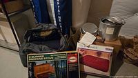 Photo showing various camping items including boxed two burner propane stove, two blue Captain's Chairs, Mirro cooking set, Cozy Rest air mattress box, and two sleeping bags.