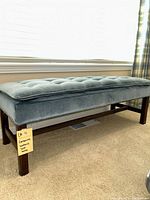 Full view of tufted blue upholstered bench on dark wooden legs in front of window with blinds and plaid curtain