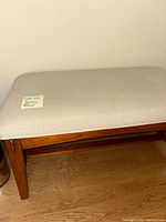 Side and partial front view of wood framed bench with light beige cloth upholstered seat on wooden floor against light wall.