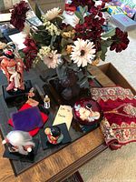 Top view showing various Asian small statues, dried flower arrangement in glass vase, decorative floral bowl, and patterned fabric piece on glass coffee table.