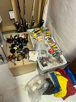 Fishing reels, tackle boxes, unopened lure packages, and raincoats in bags on the floor and box surface.