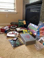 Overview photo of assortment of board games including Acquire, Denver In A Box unopened, various boxed card and board games, a 1000-piece puzzle, and other game accessories on carpeted floor near fireplace.