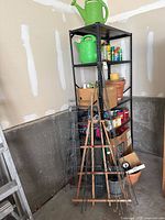 Overall view of metal shelving unit with green watering cans on top, terracotta pots, gardening chemicals, soil bags, and miscellaneous garden items around base.