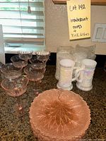 Set of six iridescent mugs, seven pink glasses, and one pink plate displayed together on counter.