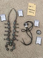 Overhead shot of squash blossom necklace, bolo tie, and circular pendant displayed on carpet with tags