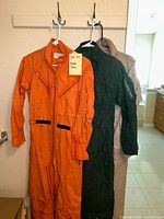 Three men’s flight suits hanging on hooks from a rack, showing orange, green, beige suits with front zip closure and pockets