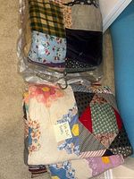 Two large patchwork quilts in folded state, one wrapped in plastic, the other exposed, showing diverse floral, plaid, and geometric fabrics