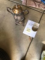 Silver plated electric percolator with cord placed on floor with handwritten note describing item and lot number.
