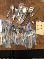 Overall view of the silverware including multiple forks, spoons, and knives, some individually bagged, along with serving pieces scattered on a wooden table.